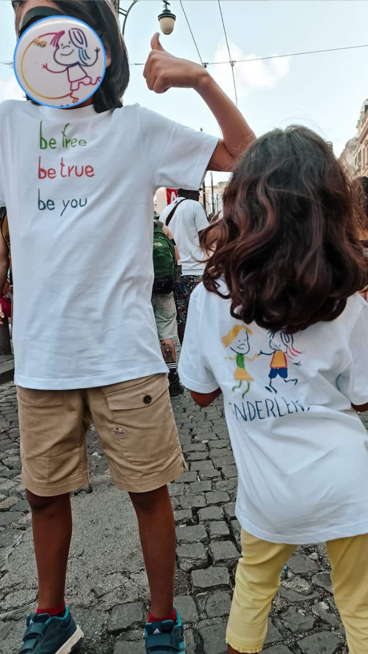 Due persone bambine al pride GenderLens: una con viso censurato sorride pollice alzato indossando maglietta "Be free be true be you", l'altra di spalle con pantaloni gialli e maglietta "GenderLens".