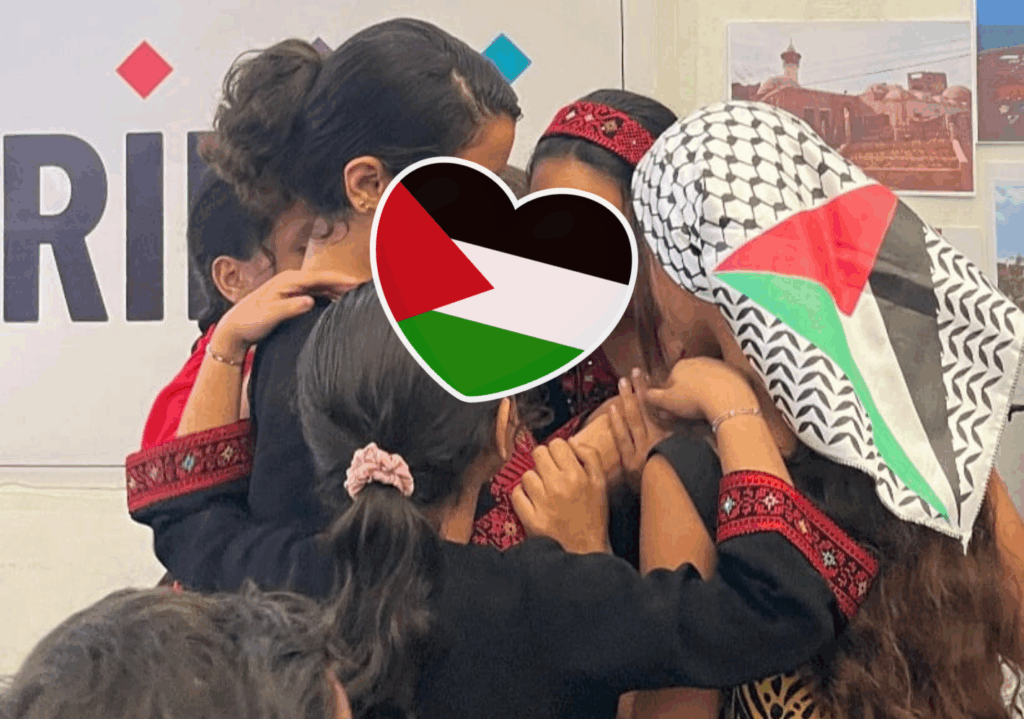 Persone bambine palestinesi in tradizionali abiti ricamati si abbracciano strette, un cuore con bandiera palestinese censura i volti.
