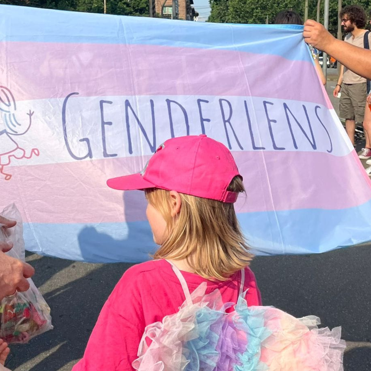 Persona bambina bionda con cappellino e maglietta fucsia e ali con i colori del trans pride, di spalle. Davanti, grande striscione con banidera trans e logo GenderLens.