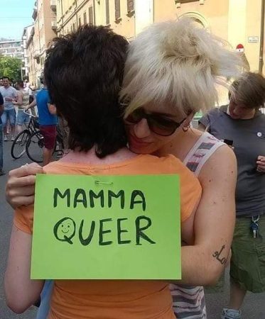 Abbraccio affettuoso tra due persone queer, una con cartello verde "Mamma queer", durante un corteo.